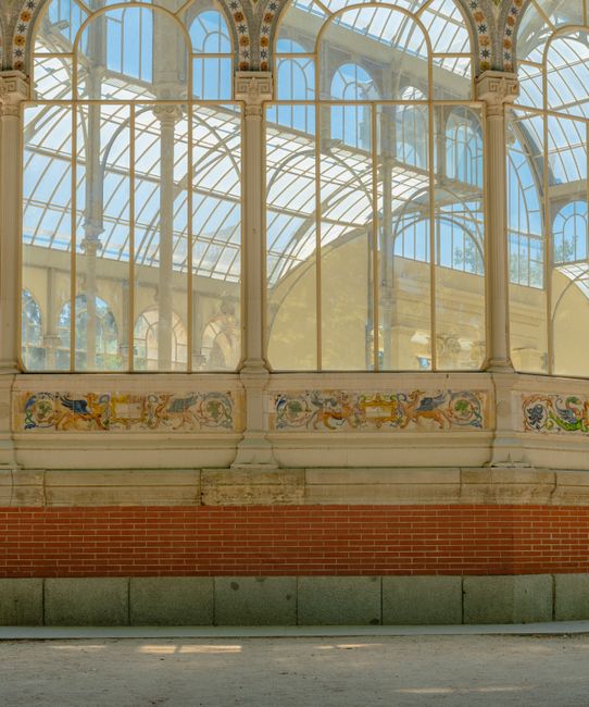 Close-up view of an ornate Victorian-style glass conservatory with decorative metal framework, showcasing large arched windows and intricate detailing. Sunlight streams through the clear panes, illumi
