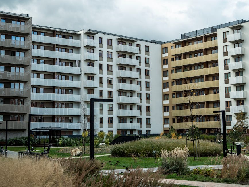 A modern residential estate featuring multiple multi-storey apartment buildings with balconies, constructed from materials including wood, glass, and concrete. The buildings are arranged around a land