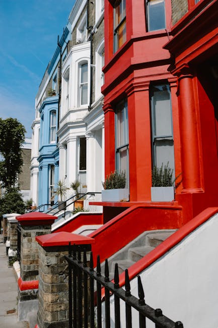 A row of Victorian-style terraced houses with stone facades, large sash windows, and gabled roofs, set against a bright blue sky with scattered clouds. The front gardens are well-maintained with a gra
